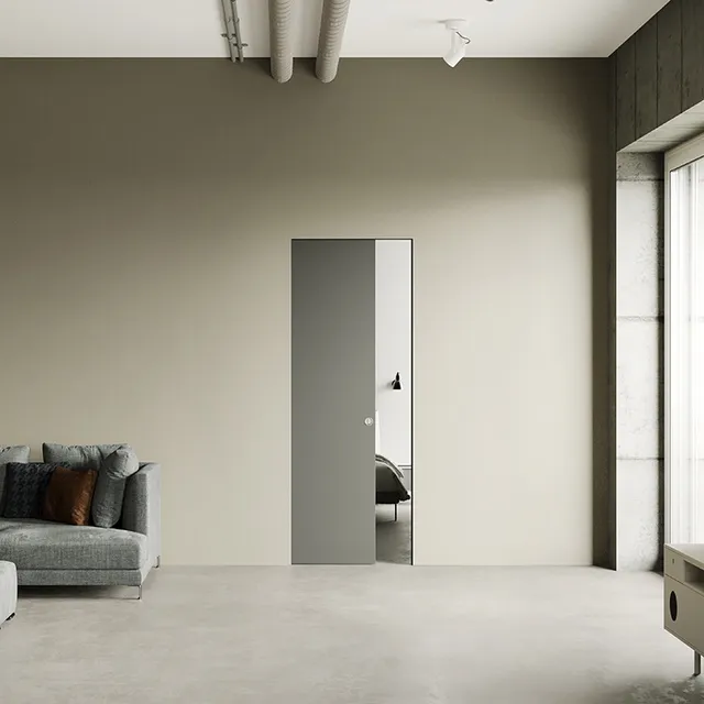 Porta scorrevole progettata per scomparire nella parete, ottimizzando lo spazio e aggiungendo un tocco contemporaneo all'ambiente. La porta è di colore grigio freddo e si presenta semiaperta. Integrata perfettamente nel muro di colore tortora.