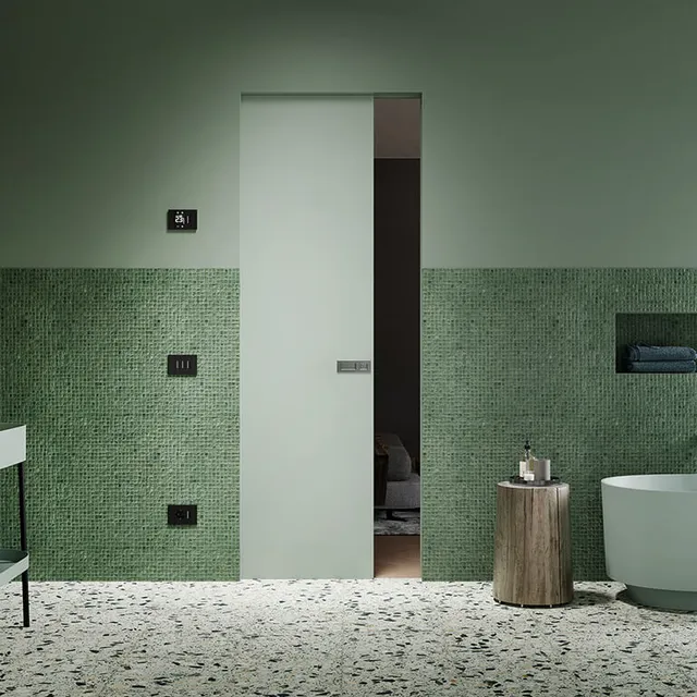 Porta scorrevole di colore verde salvia nel contesto di un bagno. Il muro in cui è integrata la porta è per metà costituito da mosaico verde acqua scuro e per metà tinteggiato in verde pastello.
